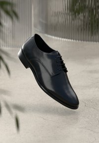 Schwarze Lederdressschuhe mit poliertem Finish, spitzer Zehenform, Schnürdesign und schlanker Silhouette. Über einer Betontfläche positioniert.