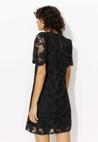 Robe en dentelle noire avec des manches courtes, motif floral, superposition texturée et un détail en forme d'ouvre-keyhole au dos, avec un ourlet légèrement évasé.