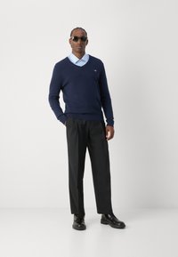 Sweater navy avec col en V, en tissu léger ; chemise à col bleu clair en dessous. Pantalon noir et boots. Modèle portant des lunettes de soleil.