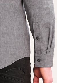 Grå langærmet skjorte med en tekstureret stof, der har knapmanchetter, et spidst manchetdesign og en buet bund. Sorte jeans synlige.