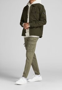 Mann in olivgrüner Jacke und Cargo-Hose, mit einer cremefarbigen Kapuzenjacke und weißen Sneakers, steht mit gefalteten Händen vor sich.