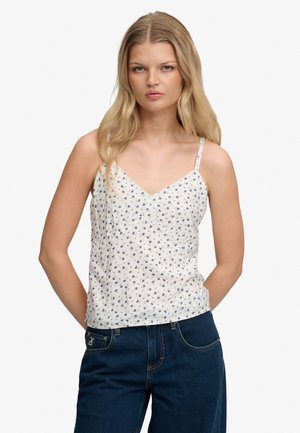 Femme aux longs cheveux blonds portant un haut blanc sans manches à fleurs et un jean bleu, debout les mains dans le dos.