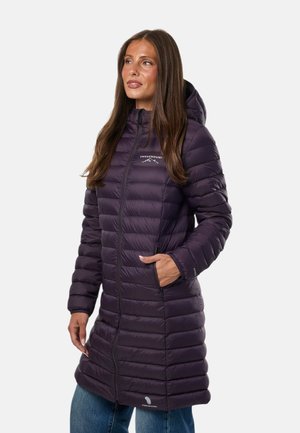 Femme portant une longue doudoune violet foncé avec capuche, debout avec une main dans la poche, légèrement tournée vers la gauche.