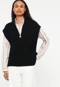 Colete de malha preto com mangas curtas e colarinho com fecho éclair, usado sobre uma blusa riscada. O material é texturado e volumoso.