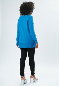 Blauer langärmliger Pullover mit geradem Schnitt, kombiniert mit schwarzen Leggings und silbernen Plateausandalen. Glatte Textur, minimale Details.