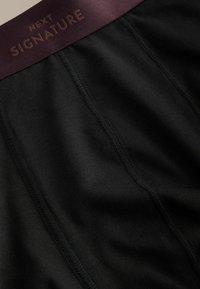 Čierna látka s vertikálnymi švami a tmavohnedý pás označený nápisom "NEXT SIGNATURE" zvýrazneným vystúpenými písmenami.