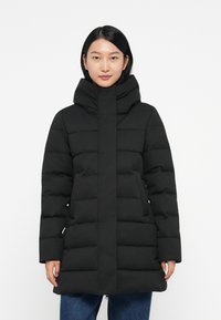 Cappotto imbottito nero con colletto alto, caratterizzato da uno stile imbottito, una zip frontale e tasche a toppa. Indossato sopra jeans blu.