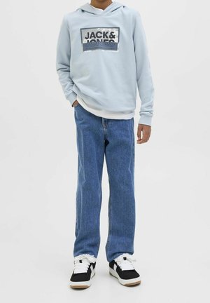Kind draagt een lichtblauwe Jack & Jones hoodie, blauwe spijkerbroek en zwart-witte sneakers, staat met één hand in de zak.