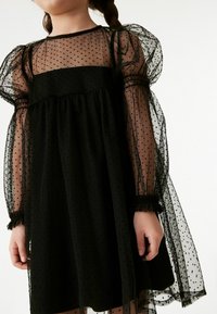 Schwarzes Kleid mit transparenten, gepunkteten Tüllärmeln mit elastischen Manschetten und einem figurbetonten Oberteil. Der Rock ist locker und fließend und erzeugt einen geschichteten Effekt.