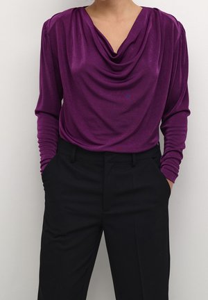 Vrouw draagt een paarse langemouwen gedrapeerde blouse, ingestopt in zwarte broek met hoge taille, handen in de zakken.