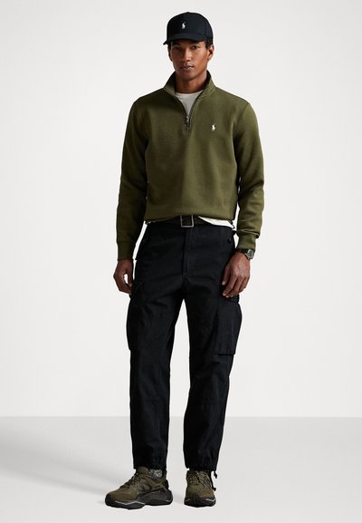 Olive green quarter-zip sweatshirt, black cargo pants, and dark trainers. Accessories include a black cap and a watch. Simple design, functional fit.