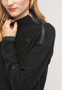 Pullover in pile nero con colletto alto, dotato di chiusura a zip e una texture a contrasto sulle spalle e sui polsini. Branding discreto sulla manica.