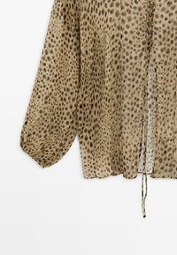 Długa beżowa bluzka z nieregularnymi ciemnobrązowymi plamami, przezroczysta tkanina, długie bufiaste rękawy, zapinana z przodu na guziki z wiszącymi troczkami.