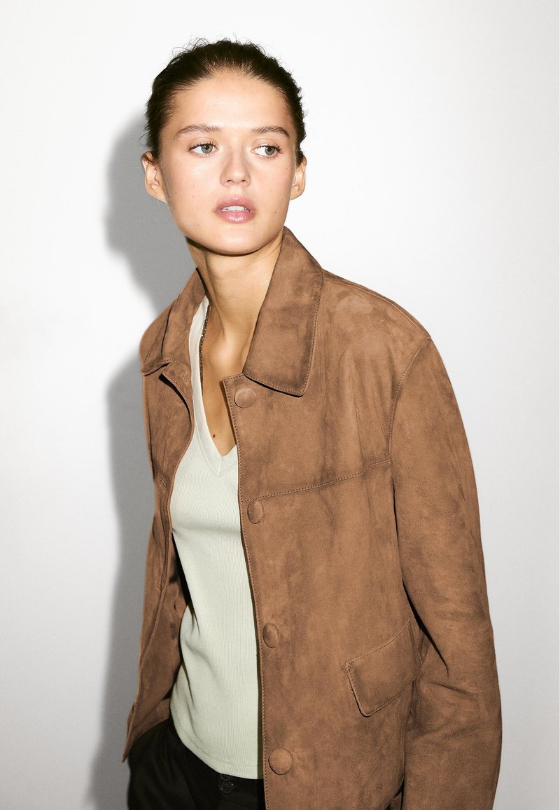 Junge Frau mit zurückgepulltem Haar, die eine braune Wildlederjacke über einem hellgrünen Oberteil trägt und seitlich vor einem schlichten weißen Hintergrund schaut.