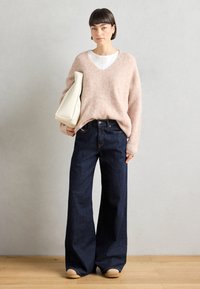Hellrosa, gestrickter Pullover mit V-Ausschnitt, kombiniert mit dunkelblauen, weitem Jeans. Das Modell hält eine cremefarbene Tasche, die eine glatte Textur aufweist.