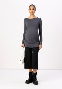 Top de maternidad gris de manga larga con fruncidos a los lados, combinado con pantalones cortos ajustados negros de canalé y botas negras gruesas con cordones, sosteniendo un bolso blanco.
