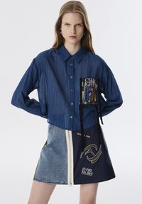 Denimblauwe overhemd met knopen en een zak waarop een grafische print staat. Gecombineerd met eenrokken die half denim en half marineblauw is, met een rits en geborduurde accenten.