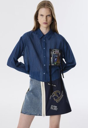 Camisa de mezclilla azul con botones y un bolsillo que muestra un gráfico. Combinada con una falda mitad de mezclilla, mitad azul marino que presenta una cremallera y detalles bordados.