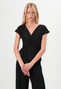 Must mustriga topp lühikeste varrukatega ja sügava V-kaelusega. Eesolev sõlm detail toob esile vöökoha. Sile, veniv kangas; ei ole nähtavaid mustreid.