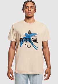Bežinis medvilninis marškinėliai su mėlynu paukščio piešiniu, juodu tekstu "PORTUGALIJA" ir dekoratyviniais elementais, tokiais kaip karūnos ir skydai.