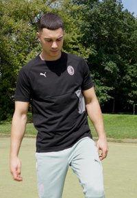 Camiseta deportiva negra de Puma con un acento gris y logo, combinada con pantalones jogger verdes claros. El entorno es un campo al aire libre rodeado de árboles.