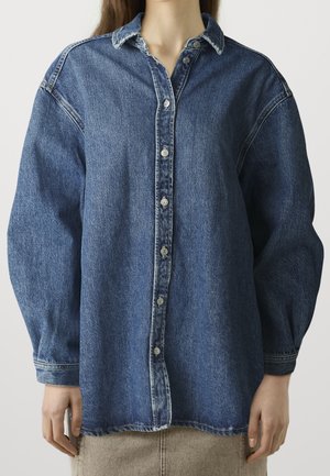 Kobieta nosząca luźną, zapinaną na guziki, długorękawową, niebieską dżinsową koszulę z opuszczonymi ramionami i beżową spódnicę na jednolitym tle.