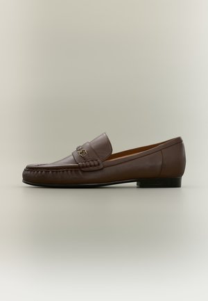 Mocasín de cuero marrón con detalle metálico en la parte delantera y un tacón negro bajo, mostrado de lado sobre un fondo neutro.