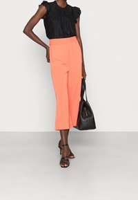 Blusa nera con ruches, pantaloni arancioni larghi, tacchi neri con cinturini, mentre tiene una borsa nera. Tessuto liscio, lunghezza alla caviglia, fascia elastica in vita.