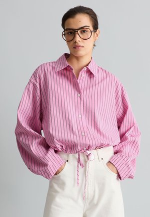 Young person wearing large glasses, a pink long-sleeve button-up shirt with black stripes, tied at the waist, and white pants, hands in pockets.