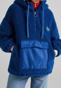 Blå fleecehoodie med en texturerad, fluffig yta, halvdragkedja, justerbara snörningar och en blå nylonficka.