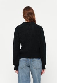Zwarte gebreide trui met een gestructureerd patroon, wijde mouwen en een ronde halslijn, gecombineerd met lichtblauwe jeans met een relaxed fit.
