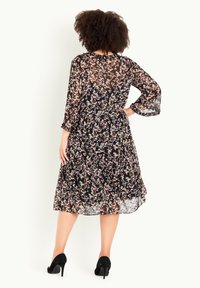 Femme aux cheveux bouclés portant une robe fleurie noire transparente à manches 3/4 et des talons hauts noirs, debout, une main sur la hanche, dos face à la caméra.