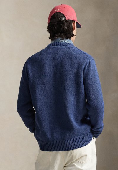 Marineblauer Strickpullover mit Struktur und geripptem Saum, getragen über einem blau gemusterten Hemd, kombiniert mit einer lachsfarbenen Mütze. Rückansicht zeigt Details und Passform.