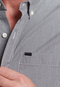 Hemd mit Knopfleiste im schwarz-weißen Karierten Muster, mit einer Tasche und einem kleinen schwarzen Markenschild. Der Stoff ist strukturiert und gewebt.