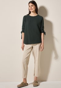 Top texturizado verde oscuro con mangas enrolladas, combinado con pantalones beige claros y zapatos marrón sin hebilla. Diseño simple y de corte relajado.
