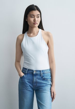 Ung kvinde med langt sort hår iført hvid ærmeløs top og blå jeans, står med den ene hånd i lommen og kigger til venstre.