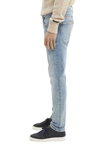 Jeans de mezclilla azul claro con un corte slim, diseño de cinco bolsillos y desvanecimiento sutil. Combinados con zapatillas oscuras y un suéter de punto beige.