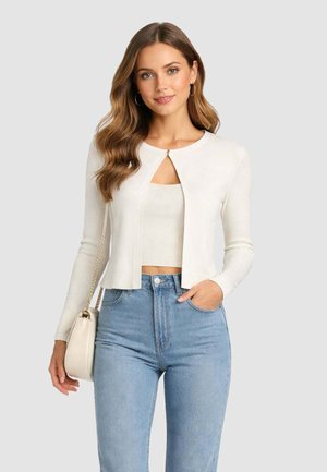 Donna con capelli castani ondulati che indossa un cardigan corto bianco sopra una maglia abbinata, jeans azzurri a vita alta e porta una borsa crema.