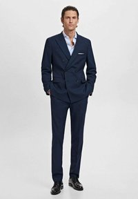 Navy double-breasted suit with a textured fabric, slim fit, paired with a light blue shirt and black shoes. Single chest pocket detail visible.