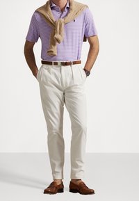 Uomo con polo a righe lavanda, maglione beige appoggiato sulle spalle, pantaloni plissettati bianchi sporchi, cintura marrone, orologio e mocassini marroni.