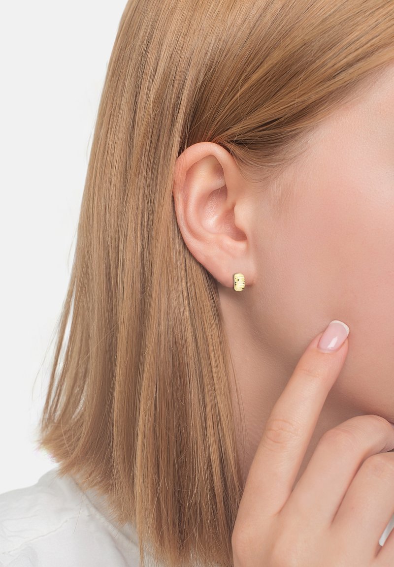 Guldstiftörhängen med en texturerad yta, med cylindrisk form och små dekorativa detaljer, bärs på en modells öra.