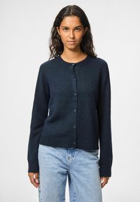 Tumši zils kardigāns ar apaļu kaklu, garām piedurknēm un pogu aizdari. Ribota apakšmala un piedurkņu gali, izgatavots no strukturētas auduma.