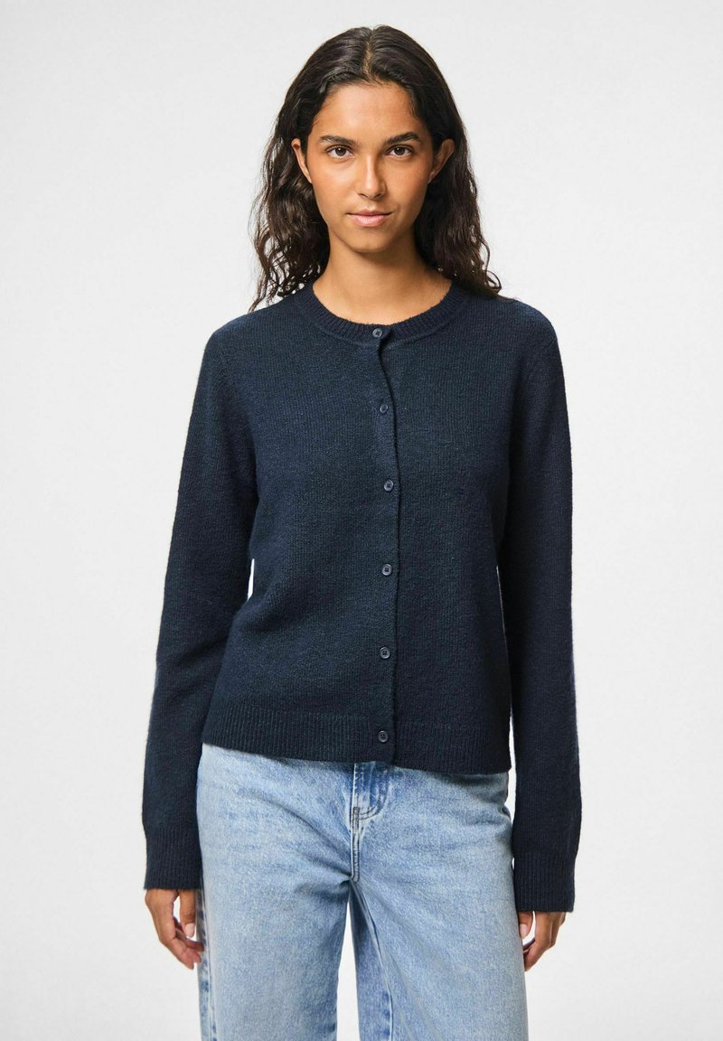 Tumši zils kardigāns ar apaļu kaklu, garām piedurknēm un pogu aizdari. Ribota apakšmala un piedurkņu gali, izgatavots no strukturētas auduma.