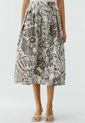 Frau trägt einen plissierten, hoch taillierten Midi-Rock mit schwarzem und weißem Blumen- und Blattmuster, kombiniert mit weißen Sandalen.