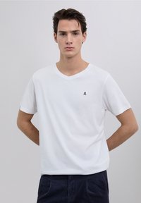 T-shirt bianca in cotone con scollo a V, presenta un piccolo logo blu navy sul petto sinistro, maniche corte e vestibilità rilassata. Texture liscia.