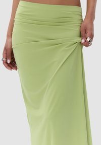 Groene, vloerlange rok van gladde stof met geplooide details bij de taille, vastgehouden aan de zijkanten door handen die ringen tonen.