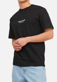 Black short-sleeve t-shirt made of cotton featuring a white "Originals Studio" logo printed on the chest. Casual fit with round neckline.