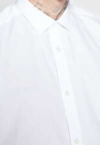 Camicia bianca button-up realizzata in tessuto leggero. Presenta un colletto classico e cinque bottoni sulla parte frontale, con una texture liscia e semplice.