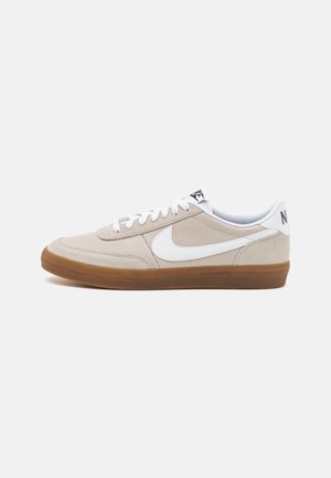 Zapatilla baja beige de Nike con el logo swoosh blanco y cordones, suela de goma marrón y logo negro de Nike en la lengüeta y talón.