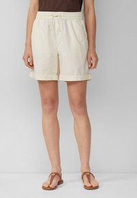 Pantalones cortos de algodón beige claro con cintura elástica, cordón ajustable y dobladillos enrollados. Llevados con sandalias de tiras marrones.
